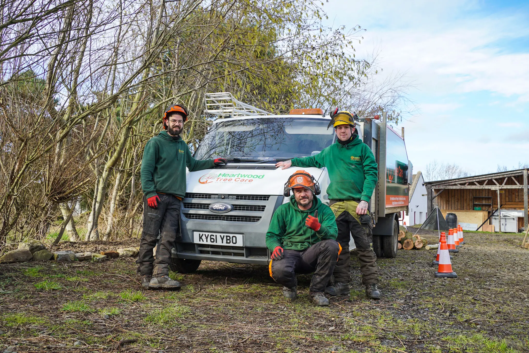 Three workers in protective gear stand and kneel in front of a truck with the "Heathwood Tree Care" logo. They are outdoors near trees, with safety cones and equipment visible. The skilled hands of these tree surgeon Edinburgh professionals ensure every job is executed safely.