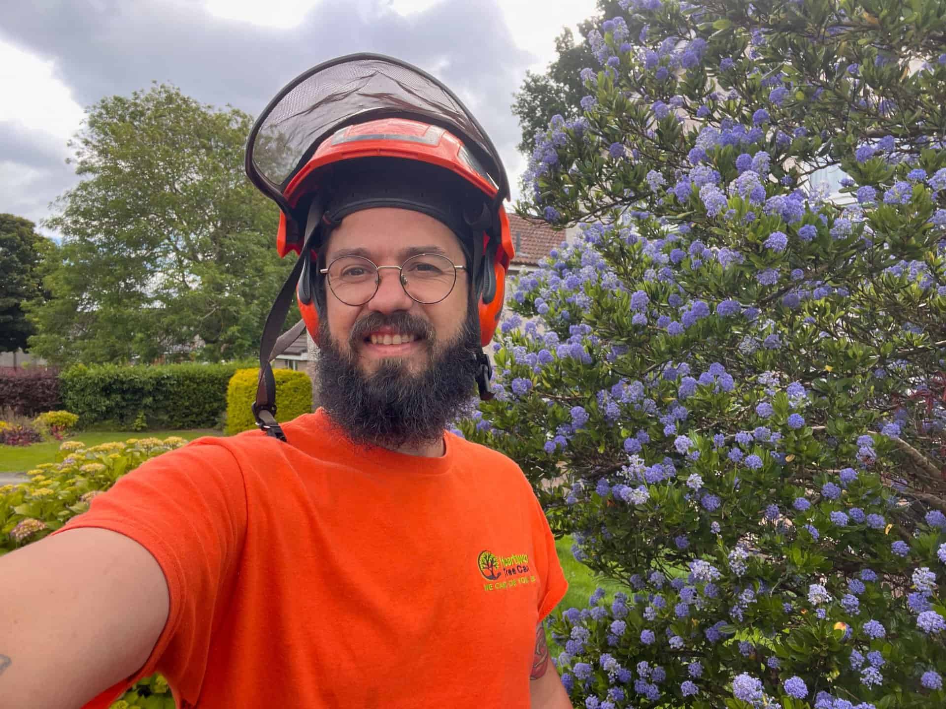 A tree surgeon from Edinburgh, wearing an orange shirt and a protective helmet with a face shield, stands outside next to a bush with purple flowers. Trees and a cloudy sky form the backdrop.
