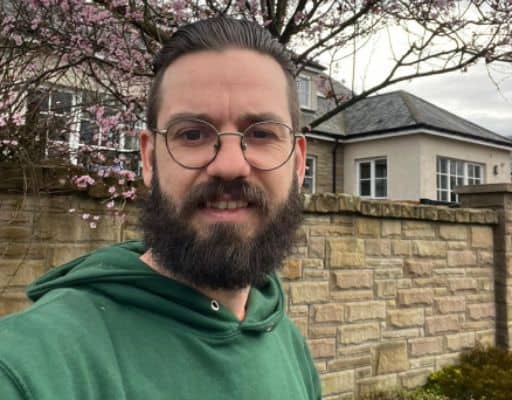 A bearded man with glasses and a green hoodie stands in front of a stone wall and a tree with pink blossoms outside a house in Edinburgh, reminiscent of the careful work of a skilled tree surgeon.