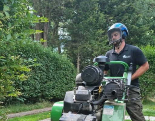 A man wearing a helmet operates a large green lawn machine near a hedge in a garden setting, much like you would see with a tree surgeon in Edinburgh.