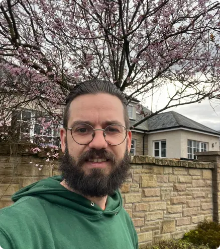 A man with glasses and a beard is standing outside in front of a stone wall and house, with a leafless tree—perhaps awaiting the care of an Edinburgh tree surgeon—and some flowering branches in the background. He is wearing a green hoodie.