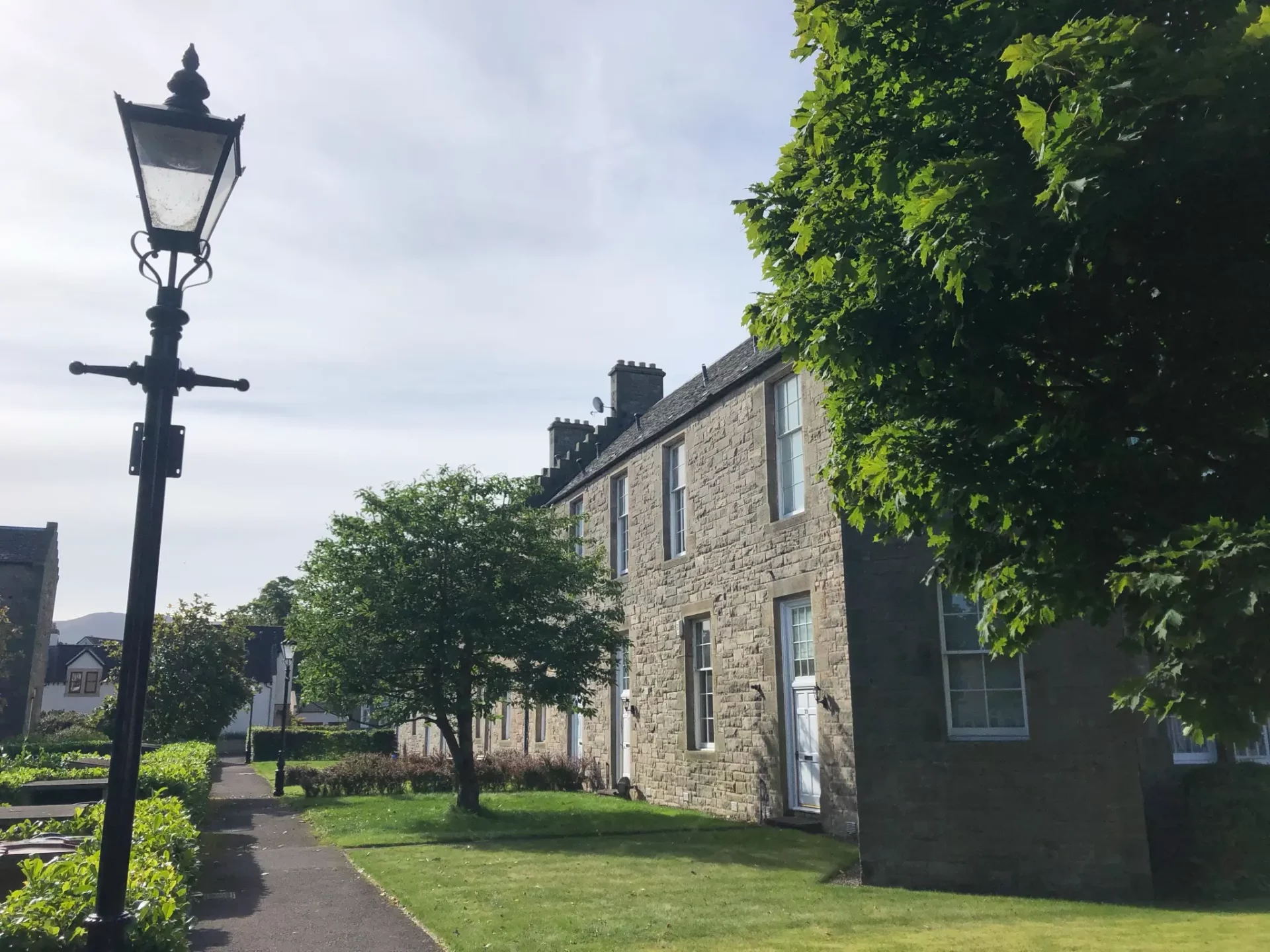 A stone building with white-framed windows and a black lamp post in front, surrounded by greenery and trees maintained by a tree surgeon in Edinburgh. A paved pathway runs alongside the house.