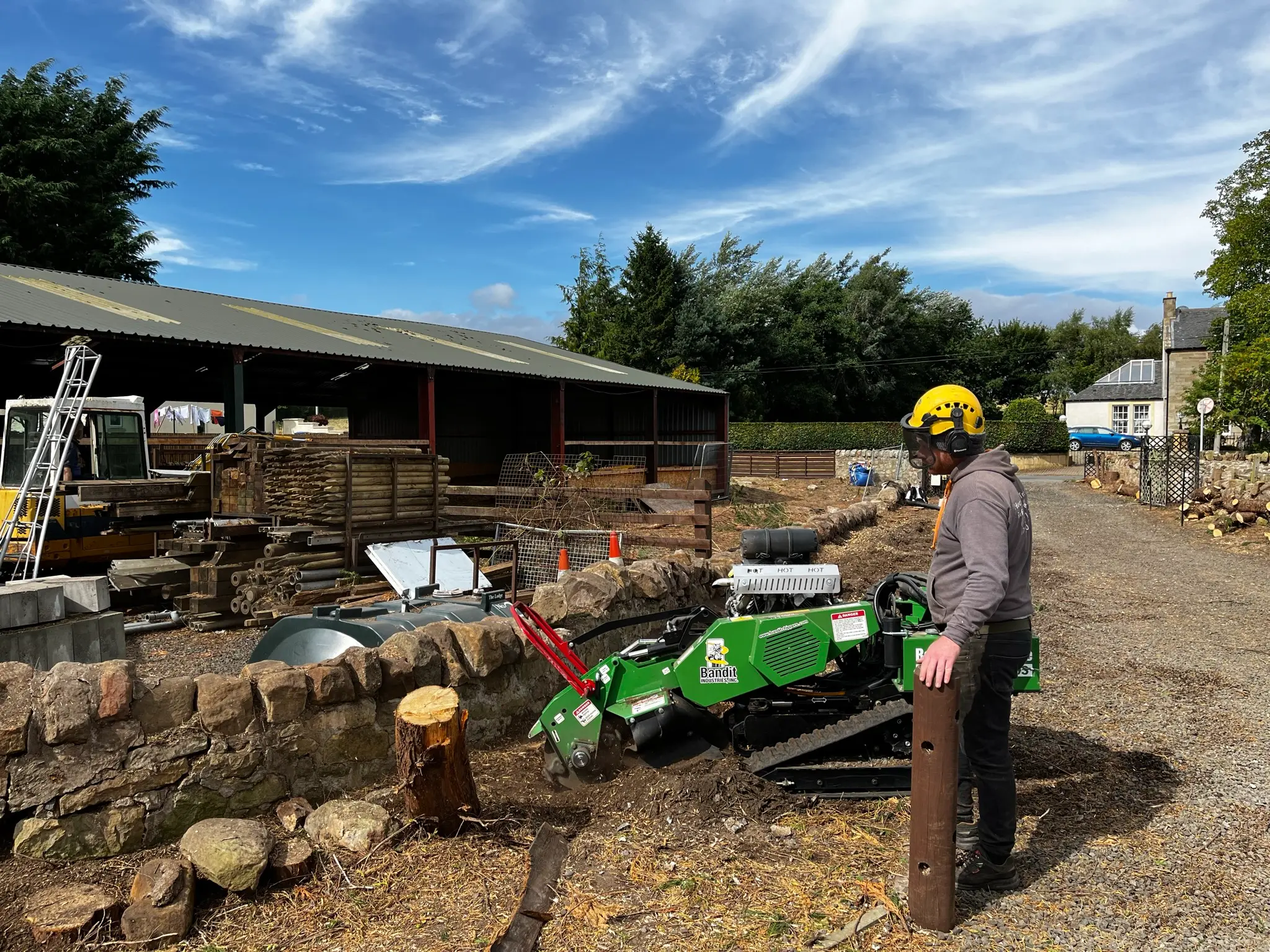 A tree surgeon in Edinburgh operates a green stump grinder near a stone fence and wooden shed on a sunny day. The person, wearing a yellow helmet and gray hoodie, efficiently clears the area.